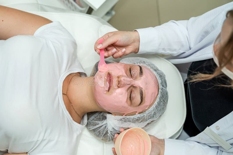 Ung kaukasisk kvinna som får ansiktsbehandling med rosa mask av beautiker vid spa salon royaltyfri fotografi