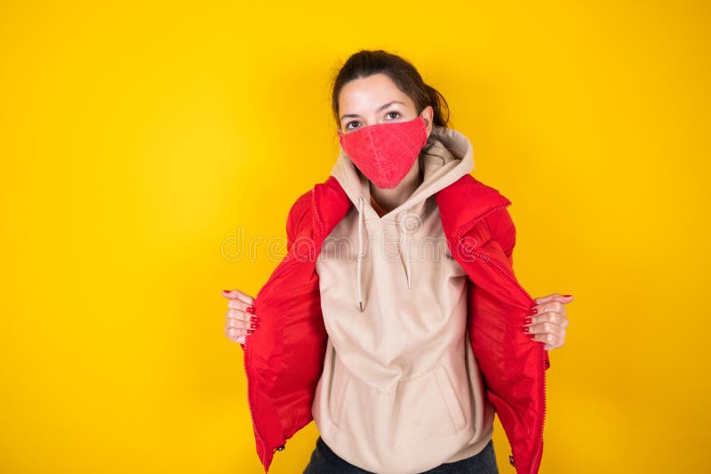 Ung kvinna i skyddande röd ansiktsmask. Kvinnor som står i hodie och jacka på gul bakgrund. royaltyfri fotografi