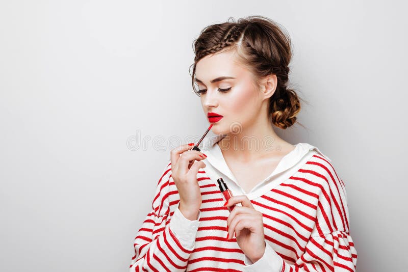 Ung kvinna med r?da kanter p? n?tt emotionell framsida i den eleganta kl?nningen som rymmer makeupl?ppstift i studio royaltyfri bild Ung kvinna med r?da kanter p? n?tt emotionell framsida i den eleganta kl?nningen som rymmer makeupl?ppstift i studio royaltyfri bild