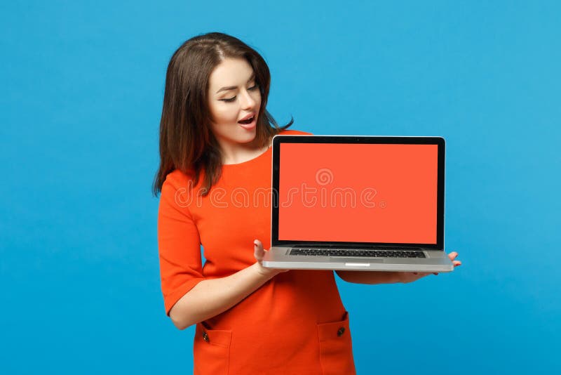 Ung kvinna med röd orange klänning i händerna med en dator för bärbar dator med tom skärm isolerad på blå fotografering för bildbyråer
