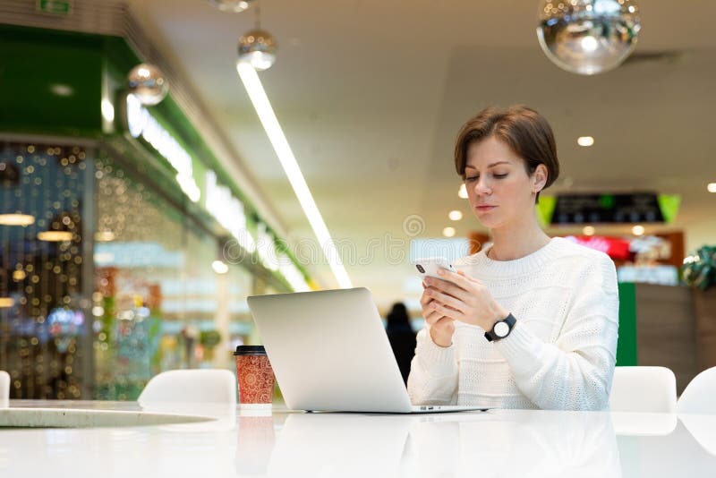 Ung kvinna som arbetar med en bärbar dator som skriver ett meddelande på telefonen arkivfoton
