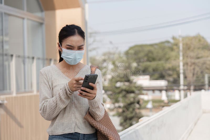 Ung kvinna som bär en mask och sms på sin smartphone utomhus i ett offentligt område. fotografering för bildbyråer