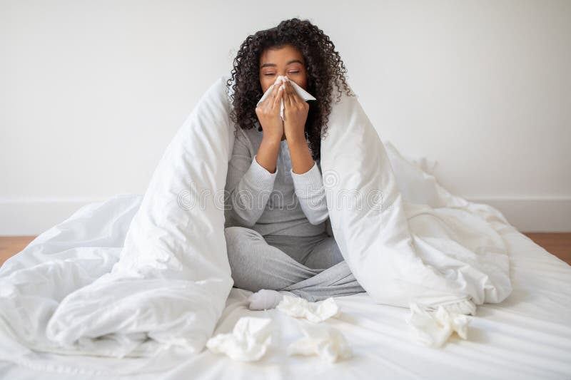 A young woman wrapped in a white duvet appears to be suffering from a cold or flu, sitting on a bed with tissues scattered around her, as she blows her nose. A young woman wrapped in a white duvet appears to be suffering from a cold or flu, sitting on a bed with tissues scattered around her, as she blows her nose