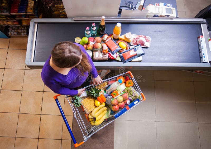 Ung kvinna som sätter gods från shoppingvagnen på räknaren för kontroll royaltyfri foto