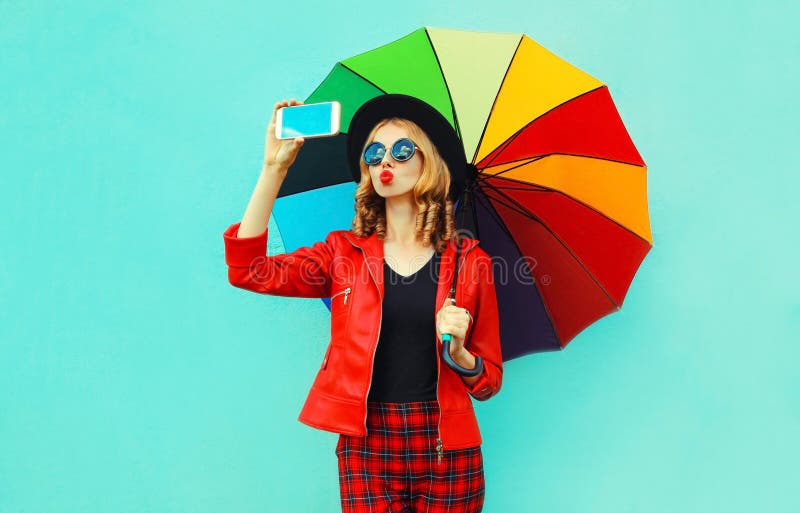 Ung kvinna som tar selfiebilden av smartphonen som blåser röda kanter som överför den söta luftkyssen med det färgrika paraplyet fotografering för bildbyråer Ung kvinna som tar selfiebilden av smartphonen som blåser röda kanter som överför den söta luftkyssen med det färgrika paraplyet fotografering för bildbyråer