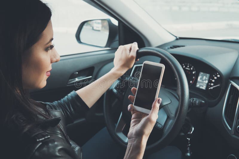 Ung kvinna som tittar pÃ¥ sin smartphone när hon kör bil i bakgrunden. Distraherad kvinnlig förare. Begrepp av royaltyfri bild Ung kvinna som tittar pÃ¥ sin smartphone när hon kör bil i bakgrunden. Distraherad kvinnlig förare. Begrepp av royaltyfri bild