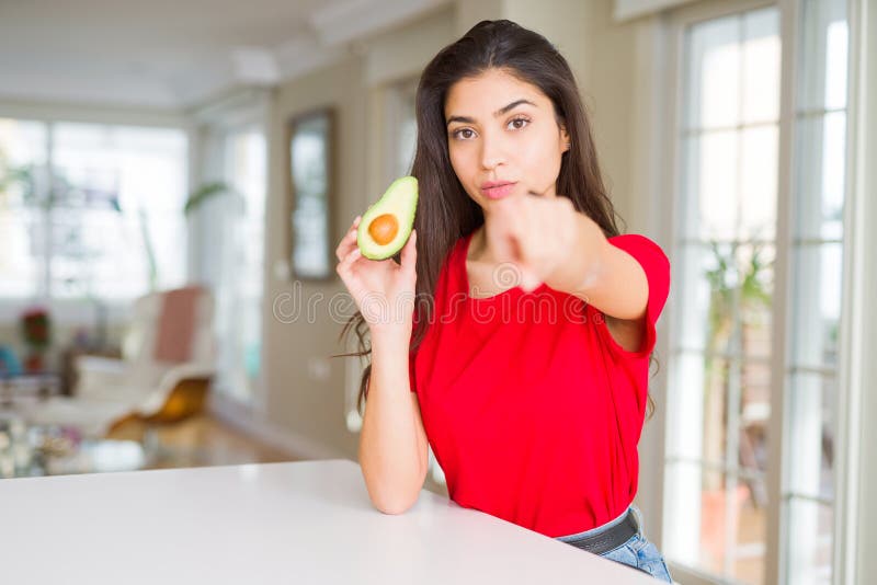Ung kvinna som äter friska avokado och pekar med fingret på kameran och till dig ger tecken på positiv och självsäker gest från royaltyfria bilder