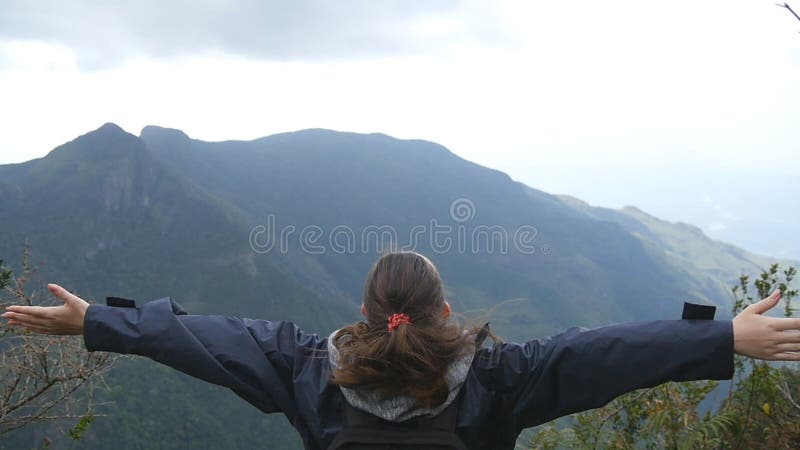 Ung kvinnlig fotvandrare med ryggsäcken som når upp överkant av berget och lyftta händer Turist- anseende för kvinna på kanten av