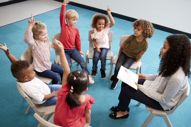 Ung kvinnlig skolalärare som läser en bok till begynnande skolbarn som sitter på stolar i en cirkel i klassrumet som lyfter hände royaltyfri bild Ung kvinnlig skolalärare som läser en bok till begynnande skolbarn som sitter på stolar i en cirkel i klassrumet som lyfter hände royaltyfri bild