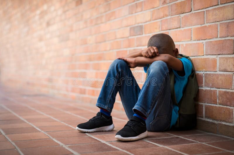 Young boy sitting alone with sad feeling at school. Depressed african child abandoned in a corridor and leaning against brick wall. Bullying, discrimination and racism concept at school with copy space. Young boy sitting alone with sad feeling at school. Depressed african child abandoned in a corridor and leaning against brick wall. Bullying, discrimination and racism concept at school with copy space.