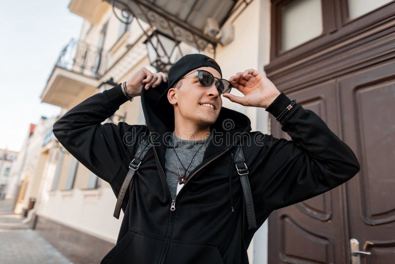 Ung lycklig hipsterman med gulligt leende i trendig svart kläder i ett lock i solglasögon med en stilfull ryggsäck i staden royaltyfri bild Ung lycklig hipsterman med gulligt leende i trendig svart kläder i ett lock i solglasögon med en stilfull ryggsäck i staden royaltyfri bild