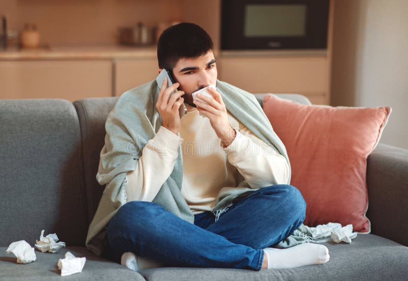 A young man, wrapped in a blanket, sits on his couch surrounded by tissues, indicating he is unwell. He holds a tissue in one hand while talking on the phone, possibly seeking medical advice. A young man, wrapped in a blanket, sits on his couch surrounded by tissues, indicating he is unwell. He holds a tissue in one hand while talking on the phone, possibly seeking medical advice