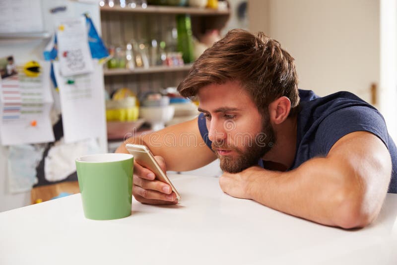 Ung man som dricker kaffe och hemma använder mobiltelefonen royaltyfri bild Ung man som dricker kaffe och hemma använder mobiltelefonen royaltyfri bild