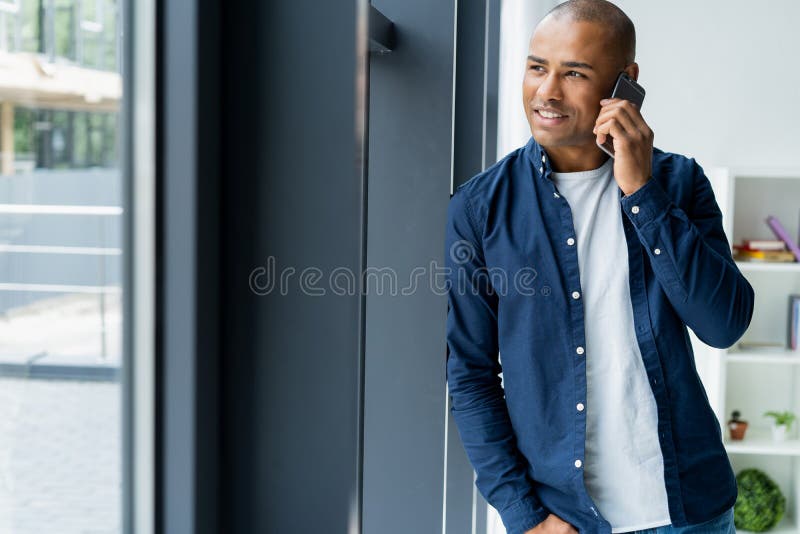 Ung man som i regeringsställning talar på hans mobiltelefon Afrikanskt utövande sammanträde på hans skrivbord med bärbara datorn royaltyfria bilder Ung man som i regeringsställning talar på hans mobiltelefon Afrikanskt utövande sammanträde på hans skrivbord med bärbara datorn royaltyfria bilder
