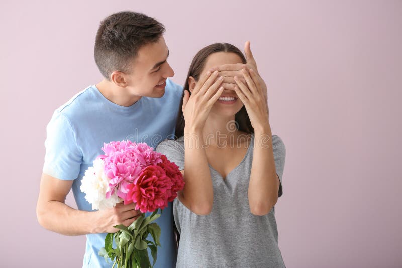 Ung man som rymmer härliga blommor och täcker ögon hans älskade flickvän på färgbakgrund royaltyfri fotografi Ung man som rymmer härliga blommor och täcker ögon hans älskade flickvän på färgbakgrund royaltyfri fotografi