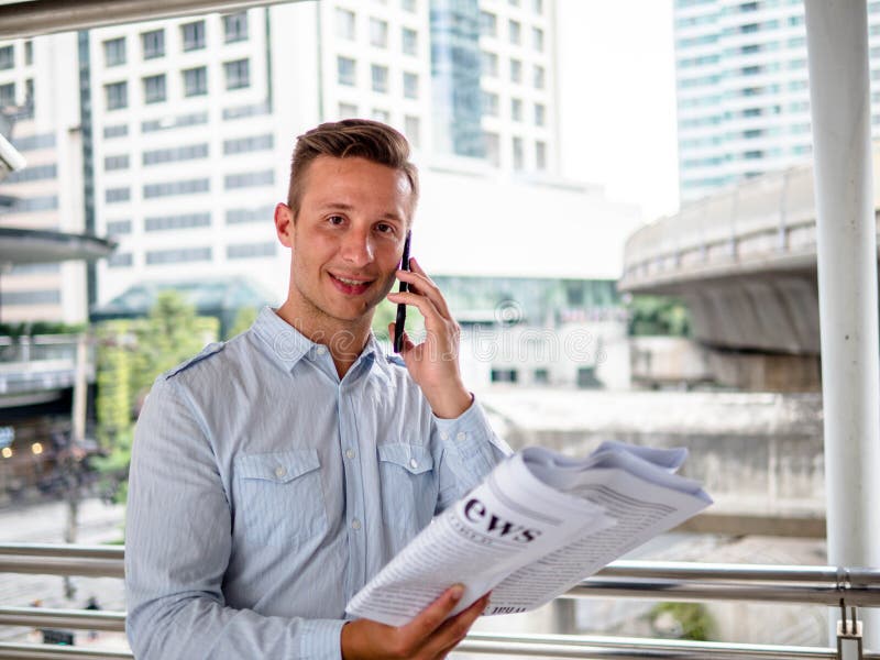 Ung man som talar på hans telefon, medan läsa tidningarna i en morgon mot bakgrunden av en modern stad royaltyfri bild Ung man som talar på hans telefon, medan läsa tidningarna i en morgon mot bakgrunden av en modern stad royaltyfri bild