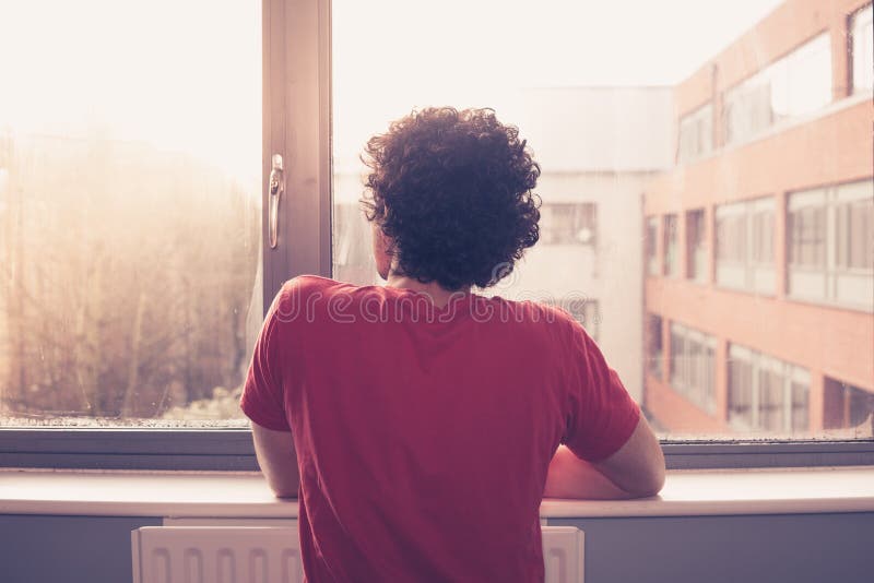 Young man is at the window silll and looking out at sunset. Young man is at the window silll and looking out at sunset