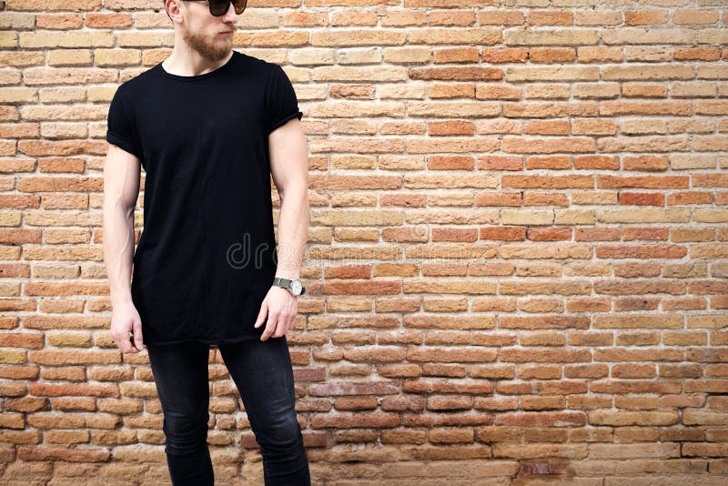 Ung muskulös man som bär den svarta tshirten, solglasögon och jeans som utanför poserar Töm den bruna grungetegelstenväggen på fotografering för bildbyråer Ung muskulös man som bär den svarta tshirten, solglasögon och jeans som utanför poserar Töm den bruna grungetegelstenväggen på fotografering för bildbyråer