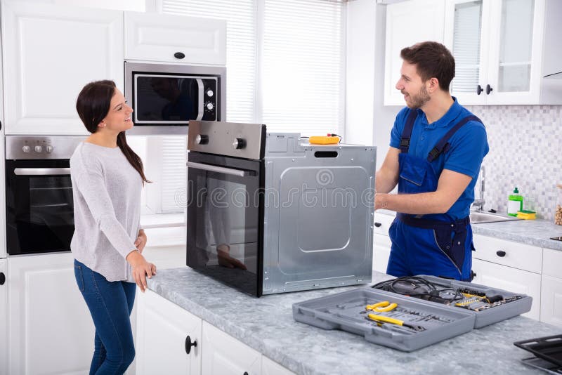Smiling Repairman Repairing Oven On Kitchen Worktop In Front Of Woman. Smiling Repairman Repairing Oven On Kitchen Worktop In Front Of Woman