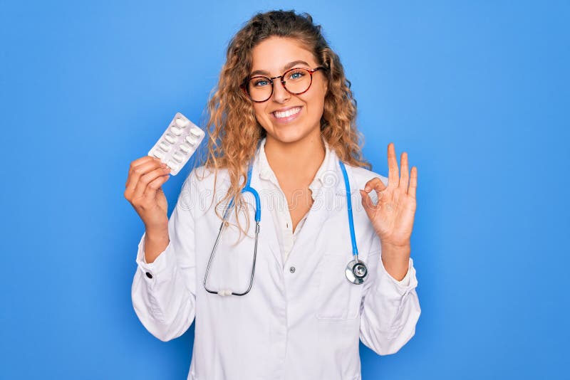 Ung, vacker blond doktor kvinna med blå ögon med stethoscope-piller som gör ok tecken med fingrar utmärkt symbol royaltyfri foto
