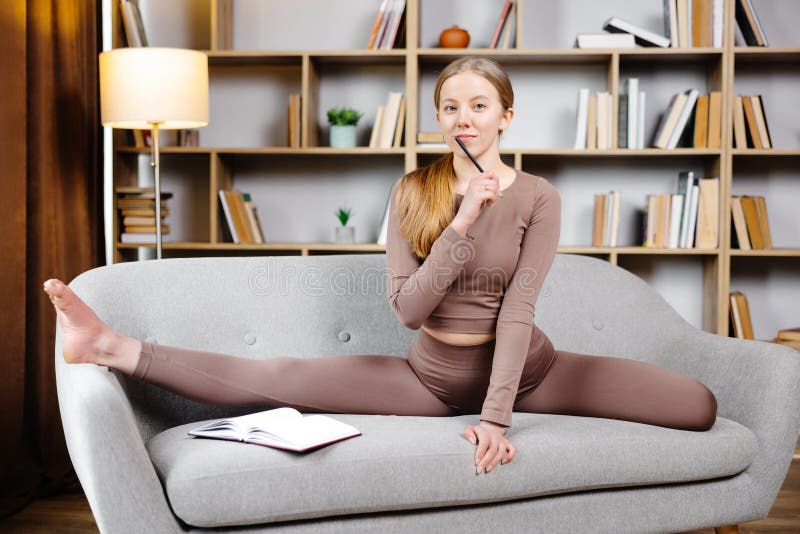 Ung, vacker blond kvinna i bruna ben och överst sittande på en tråd hemma på soffan. Flickan antecknar i en anteckningsbok arkivbilder