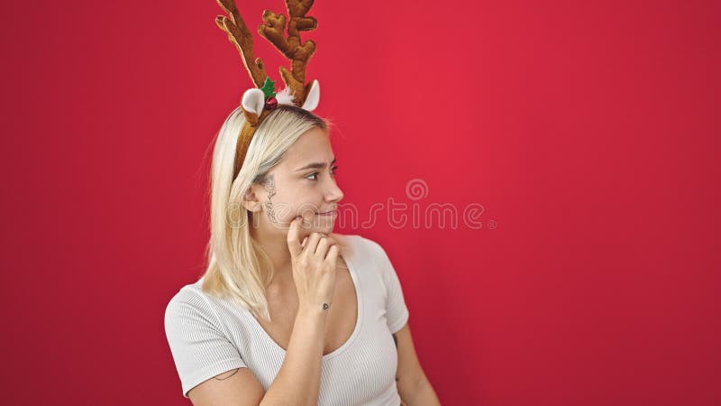 Ung, vacker historisk kvinna som bär reindeer christmas-hatt med tvivel över isolerad röd bakgrund arkivbild