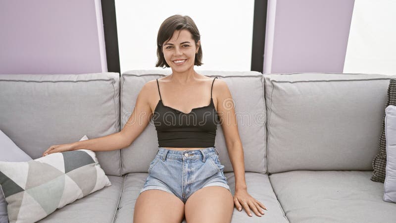Ung, vacker historisk kvinna som ler trygg och sitter på soffan hemma arkivfoto