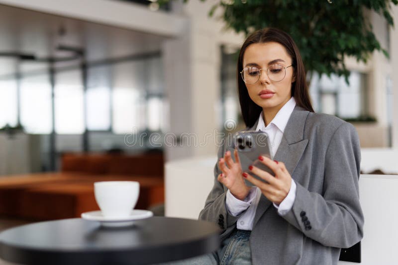 Ung vacker kvinna håller i en kaffekopp och tittar på sin smartphone medan hon sitter på ett kafé royaltyfria bilder