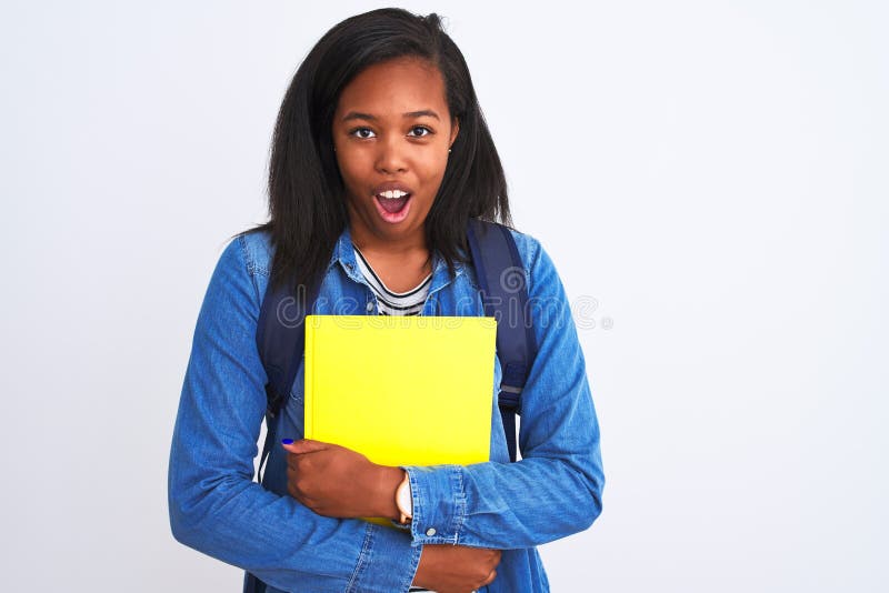 Unga afrikanska studentkvinnor som bär ryggsäck och bok över isolerad bakgrund som är skrämd av en överraskning arkivbilder