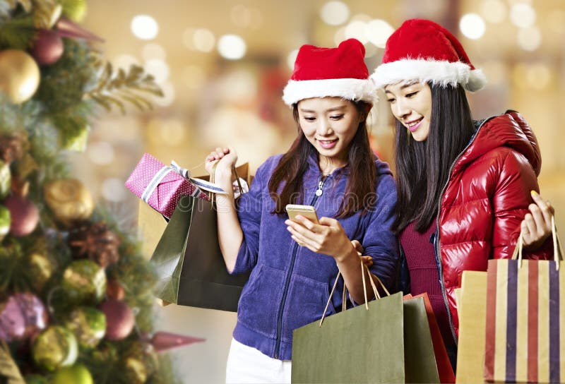 Unga asiatiska kvinnor som använder mobiltelefonen under att shoppa för jul fotografering för bildbyråer Unga asiatiska kvinnor som använder mobiltelefonen under att shoppa för jul fotografering för bildbyråer