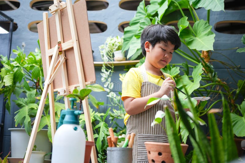 Unga asiatiska pojkmålning utomhus mellan plantor Creative Child med Easel och Brush i en grön Garden Little Artist Enjoeing arkivfoton