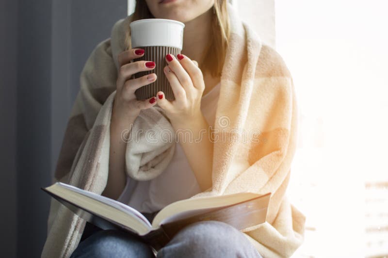 Unga flickan sitter nära fönstret som täckas med en filt, henne läser en bok och dricker te, närbild arkivbild Unga flickan sitter nära fönstret som täckas med en filt, henne läser en bok och dricker te, närbild arkivbild