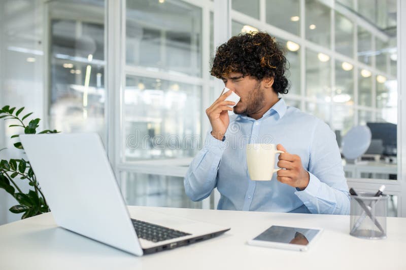 A young curly-haired man sneezing into a tissue while sipping coffee at his office desk, surrounded by modern office equipment and greenery. A young curly-haired man sneezing into a tissue while sipping coffee at his office desk, surrounded by modern office equipment and greenery.