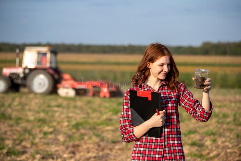 Young woman agronomist standing in field holding soil sample. Attractive young farmer monitoring land quality with tractor working behind. Young woman agronomist standing in field holding soil sample. Attractive young farmer monitoring land quality with tractor working behind