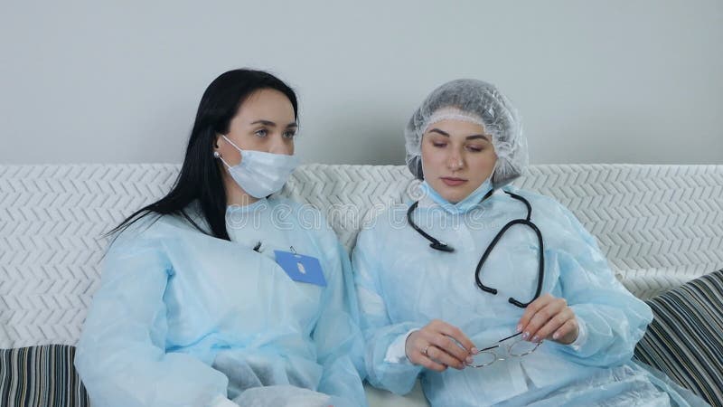Unga kvinnliga läkare med blå, jämn mask sitter och chattar på soffa efter hårt arbete stock video