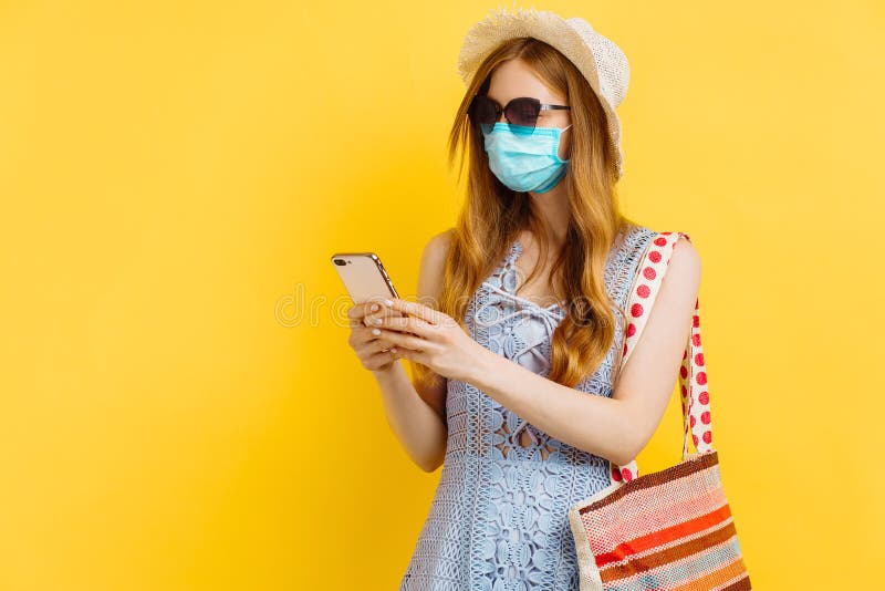 Unga kvinnor i medicinsk mask i sommarklänning och som använder mobiltelefon isolerad över gul bakgrund royaltyfria bilder