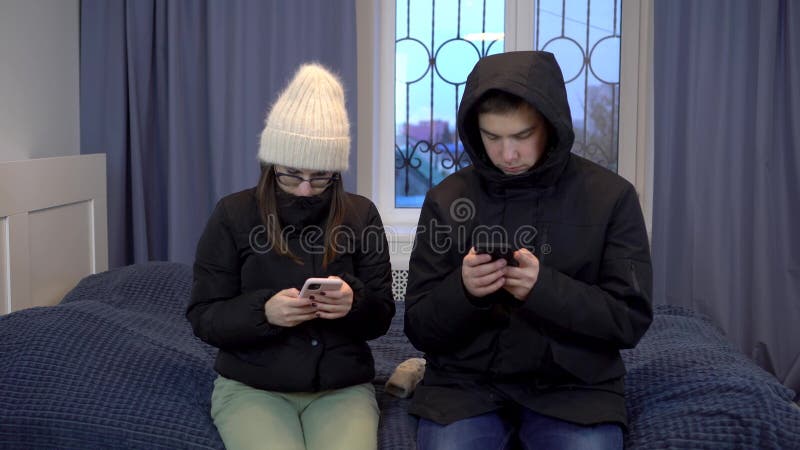 Young woman and man in jackets are sitting on the bed with phones in their hands. A young family has not paid their heating bills and is sitting in a cold apartment. 4k. Young woman and man in jackets are sitting on the bed with phones in their hands. A young family has not paid their heating bills and is sitting in a cold apartment. 4k