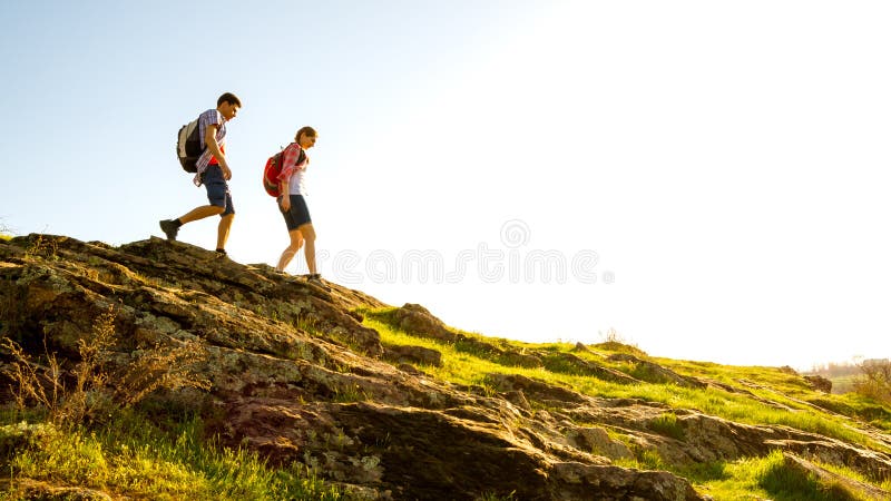Unga lyckliga par som fotvandrar med ryggsäckar på den härliga Rocky Trail på Sunny Evening Familjlopp och affärsföretag royaltyfri foto
