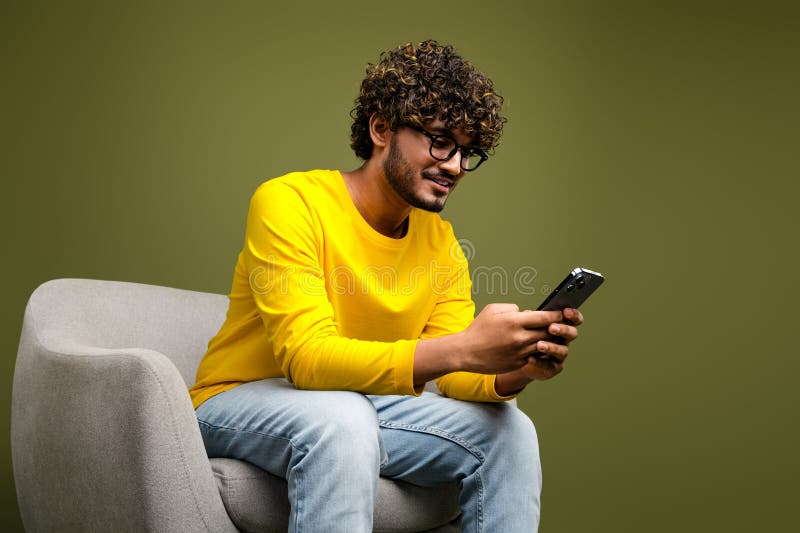 Unga man som bär en vibrant gul skjorta använder en smartphone när man sitter på en stol mot en neutral bakgrund arkivfoton