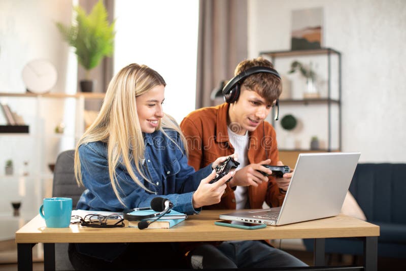 Unga nöjda par som spelar videospel hemma och sitter vid bordet med bärbara datorer och använder joysticks arkivfoto