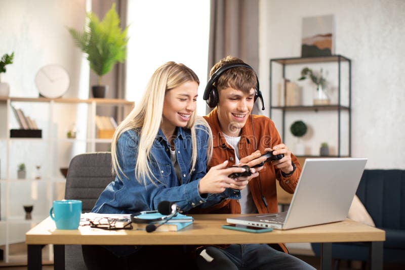 Unga nöjda par som spelar videospel hemma och sitter vid bordet med bärbara datorer och använder joysticks royaltyfria bilder