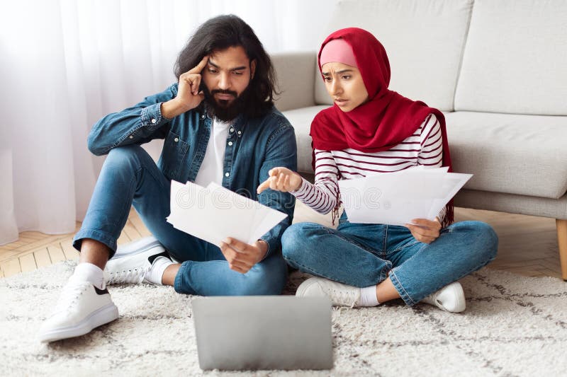 A young indian men with a beard and a women wearing a hijab sit closely on the floor with papers in hand, looking worried as they review financial documents together beside an open laptop. A young indian men with a beard and a women wearing a hijab sit closely on the floor with papers in hand, looking worried as they review financial documents together beside an open laptop