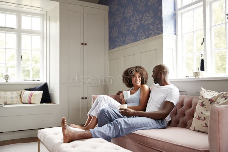 Unga svarta par som tillsammans sitter på soffan i vardagsrum som dricker kaffe och talar i morgonen, låg vinkel, full längd arkivfoto