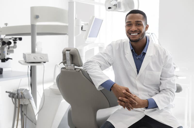 Highly qualified young black dentist posing at clinic over modern cabinet, empty space. Highly qualified young black dentist posing at clinic over modern cabinet, empty space