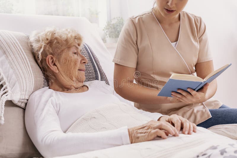 Unga vårdare läser en bok till en äldre kvinna som ligger i sjukhussängen