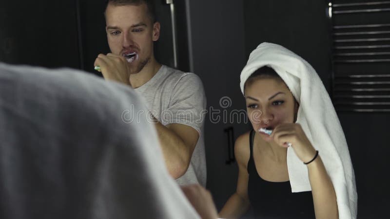 Unga älska par som ser i spegel, medan borsta tänder inomhus Bärande badlakan för dam på hennes huvud close upp