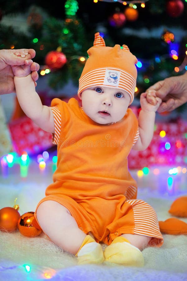 Unge i orange dräkt på en bakgrund av det julgranljusen och innehavet händerna av föräldrar royaltyfri foto Unge i orange dräkt på en bakgrund av det julgranljusen och innehavet händerna av föräldrar royaltyfri foto