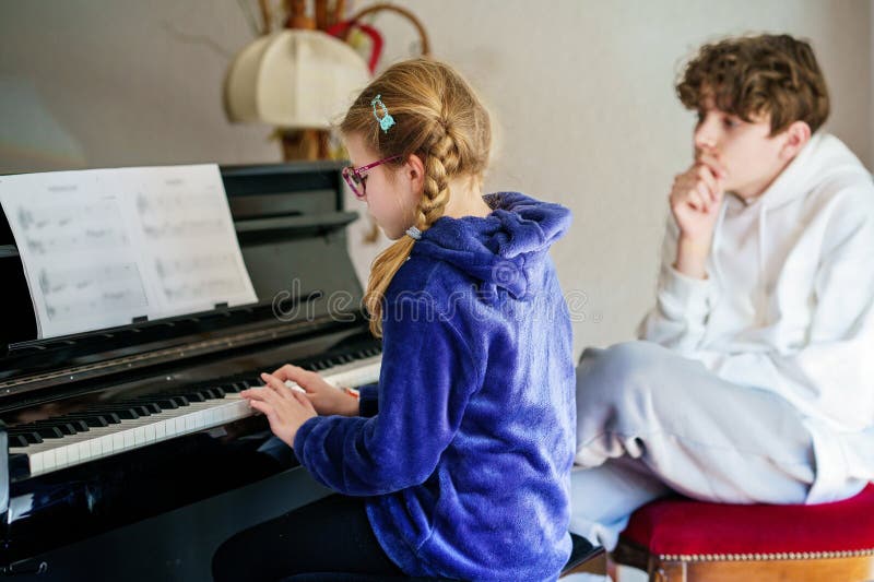 Ungefär skolflicka som utövar piano medan en tonårspojke lyssnar uppmärksamt. Barndom i musikinlärning royaltyfri fotografi