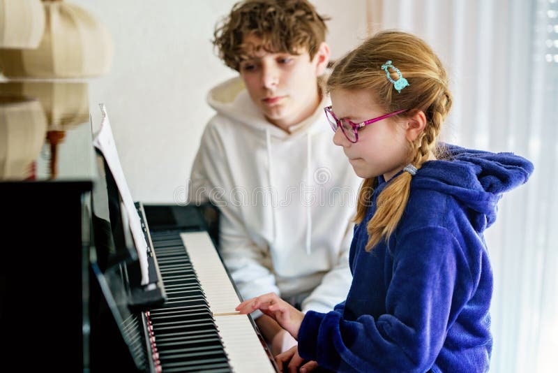Ungefär skolflicka som utövar piano medan en tonårspojke lyssnar uppmärksamt. Barndom i musikinlärning arkivbilder