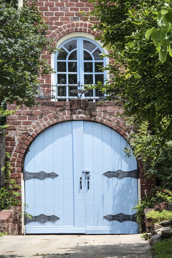 Ungewöhnlicher blauer hölzerner gewölbter Garagentor mit großen aufwändigen Scharnieren im Backsteinhaus mit kleinem Balkon mit T lizenzfreie stockbilder Ungewöhnlicher blauer hölzerner gewölbter Garagentor mit großen aufwändigen Scharnieren im Backsteinhaus mit kleinem Balkon mit T lizenzfreie stockbilder